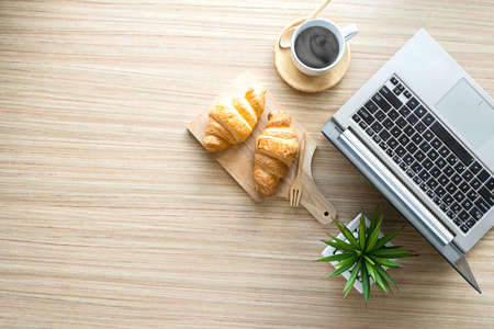 Overhead shot of modern office room with laptop computer, coffee cup, croissant on table background
