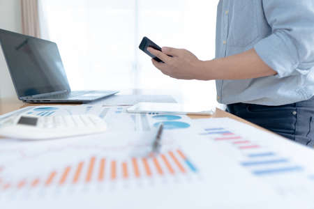 Businessman in casual wear stands beside desk to work at home with smartphone. work from home concept.の写真素材