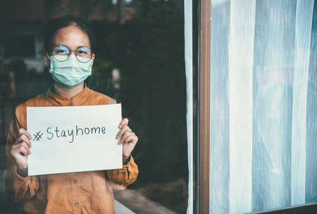 Asian girl wearing glasses, wearing a mask, holding paper labeled #stayhome, is bored of having to...