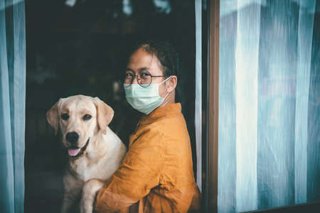 Asian girl wearing glasses, wearing a mask, with Labrador dog looking out the window, is bored of having to detain and treat the illness at home alone. Concept home quarantine, prevention COVID-19の写真素材