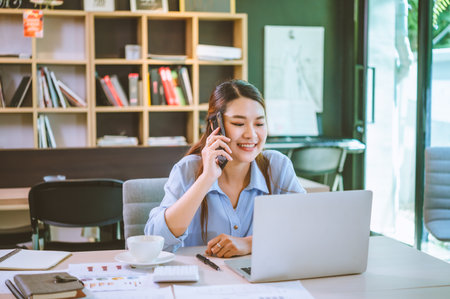 Business asian woman sitting at her desk with laptop using mobile phone chatting with customers or giving financial advice.の写真素材