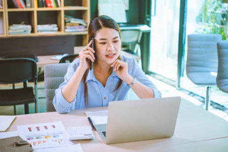 Business asian woman sitting at her desk with laptop using mobile phone chatting with customers or giving financial advice.の写真素材