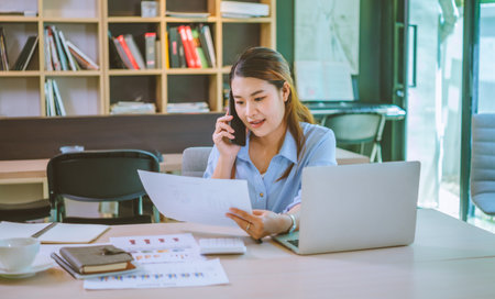 Business asian woman sitting at her desk with laptop using mobile phone chatting with customers or giving financial advice.の写真素材