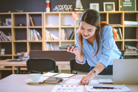 Business asian woman sitting at her desk with laptop using mobile phone chatting with customers or...