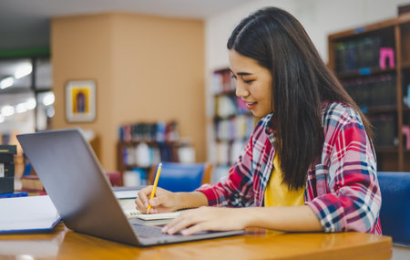 Female college student working on laptop and searching for books to study, make report, find useful...