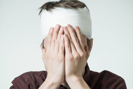 Miserable young man with bandage on his head closing face by hands. Image related with treatment of the woundsの写真素材