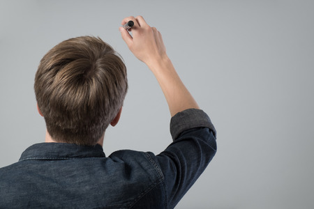 Young man with pen writing on virtual screen isolated on greyの写真素材
