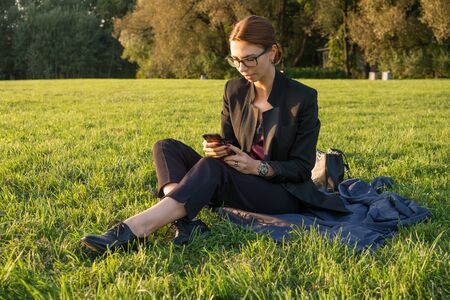 Young businesswoman in glasses using her smartphone and sitting on green grass at sunny day. Working outdoor, staying connectedの写真素材