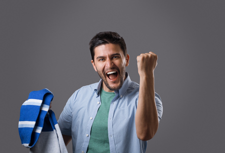 Portrait of young bearded sports fan with blue scarf holding his fists over his head isolated on grey background. Support your teamの写真素材