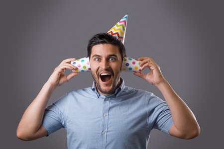 Handsome cheerful young brunet guy with party cap covering ears by paper cups. New year or birthday celebrationの写真素材