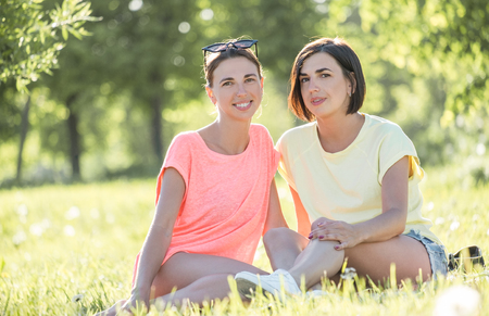 Outdoor portrait of two cheerful girls sitting on lawn and having good time together in park at sunny day.の写真素材