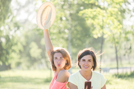 Two handsome girls, laughing sitting on lawn and listening music on smartphone together in green park at sunny warm day. Outdoor time spending in summer dayの写真素材