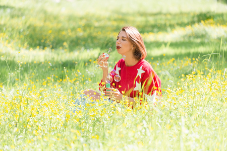 Young handsome girl sitting on grass and blowing soap bubbles in the air at sunny day in parkの写真素材