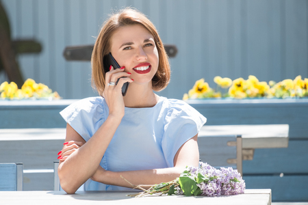 Outdoor portrait of young charming woman in light blue dress sitting at table of summer cafe and having conversation at smartphone  with bouquet of fresh lilacの写真素材