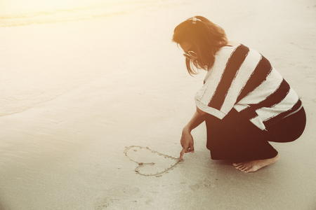 Single broken heart girl concept. Hipster alone women drawing one heart with her finger on the sea beach vintage warm color tone.の写真素材