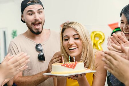 American blond beautiful girl blowing the birthday cake in the party with friends clap her birthday celebration song.の写真素材