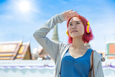 Asian girl teen red hair colored standing outdoor against high UV sun light at summer season sunny day sky Thai temple background.の写真素材