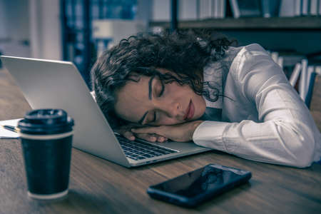 Tired working women sleeping at office desk hard work overnight nap. African American black businesswomen employee worked late and fell asleep on the computer laptop table at night with coffee cup.の写真素材