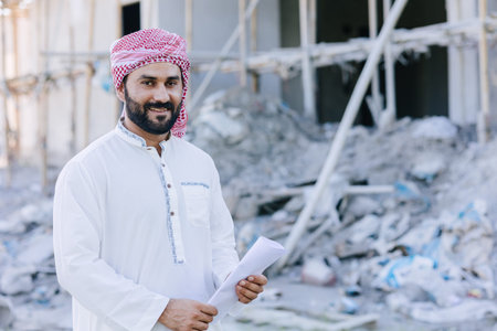 Arab muslim adult engineer business male portrait standing with floor plan at construction siteの写真素材
