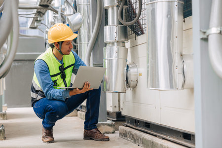 A man in a yellow vest is looking at a laptop while kneeling down. He is wearing a hard hat and safety glassesの写真素材