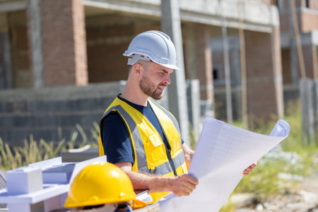 Construction worker with floorplan. Caucasian engineer male wearing safety vest and helmet at...
