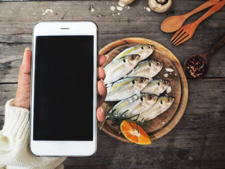 Smart phone on hand with fresh mackerel fish and spices on wood backgroundの写真素材