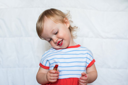 Little funny blond baby girl make up lips with mom's red lipstick. Learning to be woman. Happy...