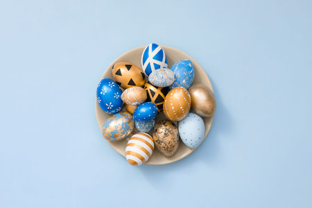 Stylish Easter golden decorated eggs on grey plate isolated on blue background. Trendy flat lay...