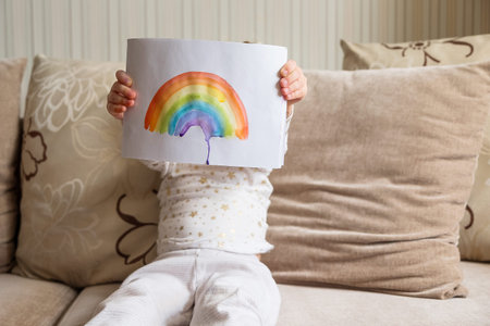 Kid painting rainbow during Covid-19 quarantine at home. Girl holding adrawing with rainbow. Stay at home Social media campaign for coronavirus, lets all be well, hope during pandemicの写真素材