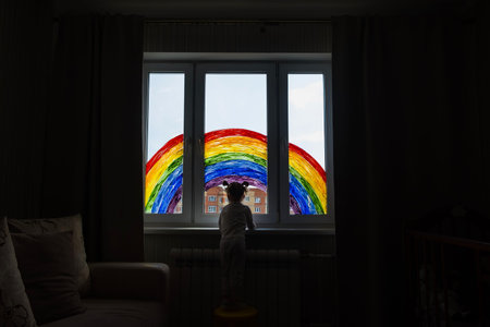 Little girl on background of painting rainbow on window. Kids leisure at home. Positive visual support during quarantine Pandemic Coronavirus Covid-19 at home.の写真素材