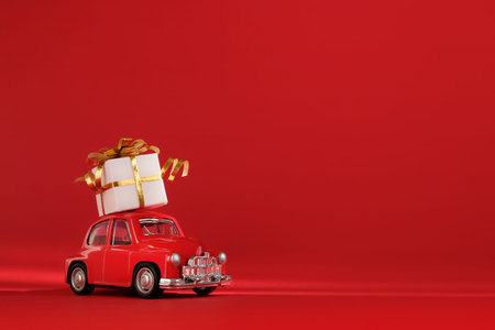 Red small retro toy car with gift on the roof on red background. Delivery. New Year, Christmas,...