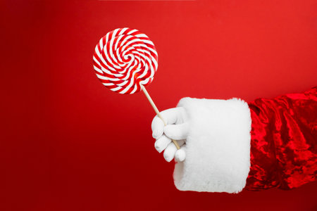 Santa hand with colorful lollipop on red background, wooden stick, red and white spiral, childhood sweets, Christmas concept.の写真素材