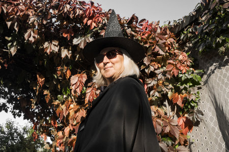 Portrait of old blonde woman in witch costume and hat for halloween and glasses on fall autumn leaves background. Happy Halloween.の写真素材