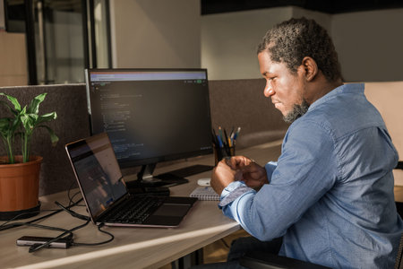 Black Man Writing Lines of Code On Desktop PC With Two Monitors and a Laptop Aside in Stylish...
