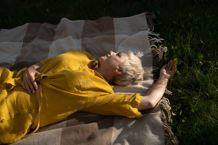 A serene scene of a mature woman dressed in yellow, leisurely relaxing on a blanket in her garden...