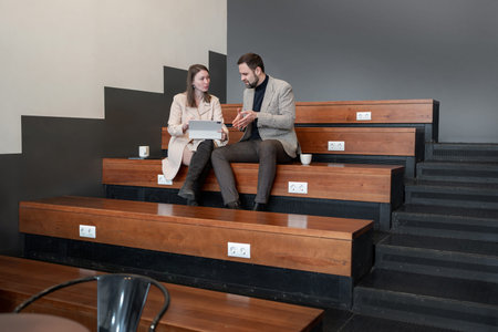 Business partners in conversation on tiered wooden seating in modern workspace. Conversation, tiered...