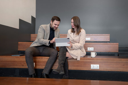 Man and woman reviewing paperwork together on stepped wooden bench in modern workspace. Paperwork...