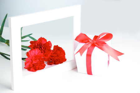 Three red carnation flowers, gift in white packaging with a red bow and frame on a white background. Greeting card for mother's day, international women's day.の写真素材