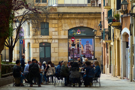 Tarragona, Spain - April 15, 2024: Urban scene in Tarragona where individuals gather for coffee, with a picturesque view of an old street and historic architecture in the background.のeditorial素材