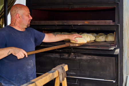 Viladecans, Barcelona, Spain-May 26, 2024: A tattooed baker handles an industrial oven to bake artisan bread, showcasing culinary skill and tradition.のeditorial素材