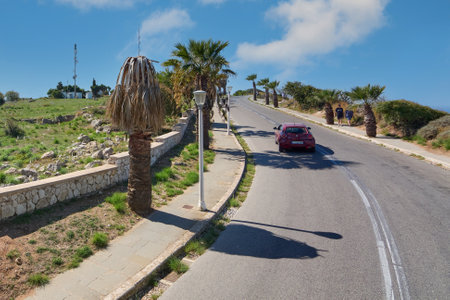 Rhodes. Greece - June 06, 2025: A road ascends in Rhodes, Greece, flanked by palm trees and a stone wall, with a red car and a blue sky, reflecting the island's charm.の写真素材