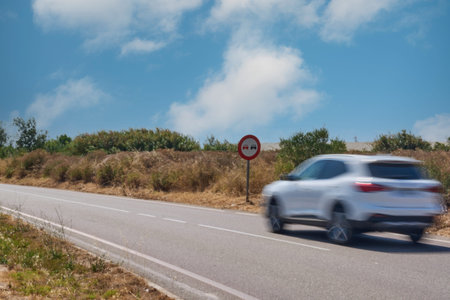 A white car is captured in motion blur as it passes a road sign on a sunny day, symbolizing traffic...