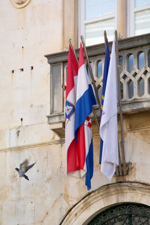 Flags of Croatia, the European Union, and the city of Dubrovnik (Saint Blaise) hang from a stone balcony in the old town. A pigeon flies past the facade.の写真素材