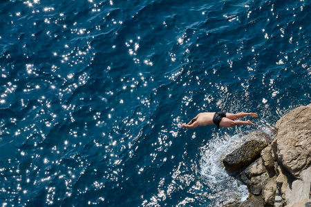 High angle shot of a man diving from a rocky cliff into the deep blue sea. The water sparkles brightly in the sun, evoking adventure and freedom.の写真素材