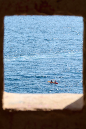 Two people paddle a tandem kayak on the sparkling Adriatic Sea. The scene is framed by an out-of-focus stone window from the Dubrovnik city walls.の写真素材