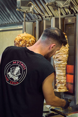 Corfu. Greece - January 26, 2026: Employee at Mythos and Grill restaurant stands near a vertical rotisserie preparing greek gyros in a professional kitchen environment during busy lunch service.のeditorial素材