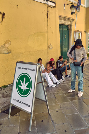 Corfu. Greece - January 29, 2026: Legal cannabis sign stands on a narrow street as young people sit and wait nearby, reflecting changing drug laws, urban youth culture, and modern social attitudes in Europe.のeditorial素材