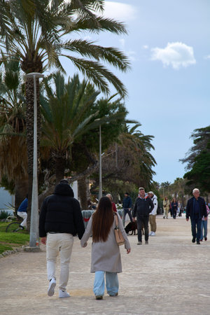 Sitges. Spain - February 12, 2026: Young couple walking hand in hand along palm tree lined seaside...