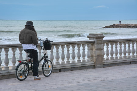 Sitges. Spain - February 15, 2026: Woman with folding bicycle checking smartphone along seaside promenade, urban commuting and digital lifestyle by the Mediterranean coastのeditorial素材