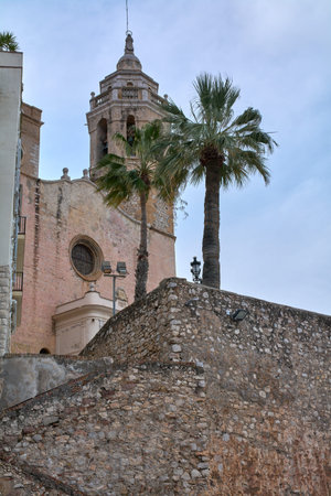 Church of Sant Bartomeu and Santa Tecla bell tower rising above historic stone wall and palm trees under overcast sky.のeditorial素材